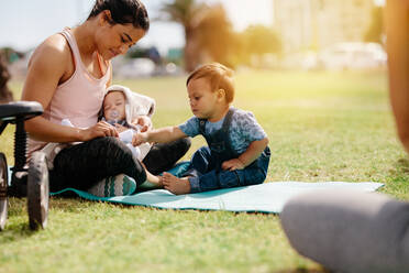 Young mom in fitness wear sitting in park with her kids. Woman sitting with her kids on grass early in the morning with sunflare in the background. - JLPSF20066