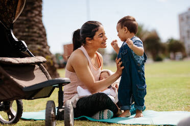 Junge Mutter sitzt mit ihren beiden Kindern morgens auf einer Yogamatte im Park. Frau entspannt sich nach ihrem Morgenspaziergang mit Kindern im Park. - JLPSF20067