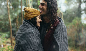 Verliebtes Paar, das in eine Decke eingewickelt im Regen steht. Lächelnder Mann und lächelnde Frau, die sich an einem regnerischen Tag im Wald umarmen. - JLPSF20073