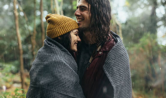 Verliebtes Paar, das in eine Decke eingewickelt im Regen steht. Lächelnder Mann und lächelnde Frau, die sich an einem regnerischen Tag im Wald umarmen. - JLPSF20073