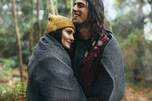 Beautiful young couple wrapped in a blanket on a rainy day. Caucasian couple embracing under the rain. - JLPSF20074