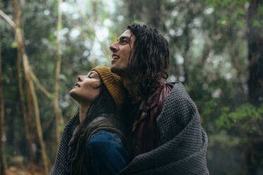 Portrait of young couple covering themselves with blanker while standing under rain in park. Beautiful couple spending time together in rain. - JLPSF20075