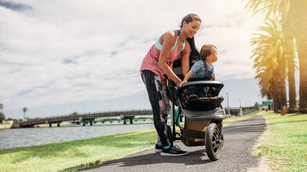 Frau mit ihrem Baby im Kinderwagen beim Morgenspaziergang. Junge Mutter mit ihrem Baby im Kinderwagen im Park am See. - JLPSF20083