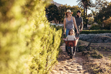 Woman pushing little girl on wheelbarrow with man walking behind. Young family enjoying in their farm. - JLPSF20086