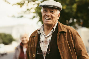 Portrait of happy senior man in warm clothes and cap looking at camera with a woman in background. Retired man standing outside on street on a winter day. - JLPSF20099