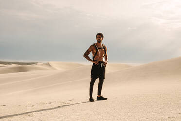 Junger muskulöser Mann in voller Länge, der auf einer Sanddüne steht und mit den Händen in den Hüften wegschaut. - JLPSF20104