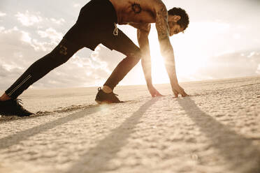 Fit young man in starting position ready for running. Athlete ready to start a race over desert sand with sun shining in background. - JLPSF20114