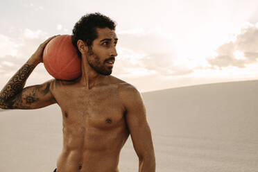 Muskulöser Mann mit einem Gymnastikball in der Wüste. Sportler mit einem Medizinball für Fitnesstraining auf Sanddünen. - JLPSF20117