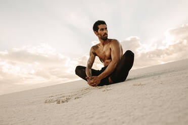 Athlete doing stretching exercises sitting on sand dune in evening. Runner sitting in desert with feet joined together and looking away. - JLPSF20121