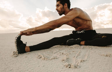 Sportler beim Stretching-Training auf einer Sanddüne sitzend. Läufer sitzt in der Wüste und dehnt die Beine. - JLPSF20122