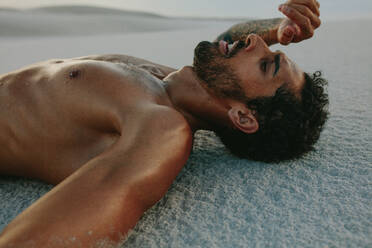 Tired man lying on sand dune after intense physical training. Athlete resting on sand after workout session. - JLPSF20127