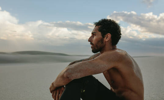 Seitenansicht eines muskulösen männlichen Athleten, der im Wüstensand sitzt und die Aussicht betrachtet. Ein müder Mann macht eine Pause vom Training in der Wüste. - JLPSF20131