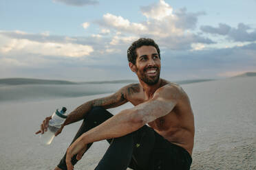 Lächelnder muskulöser männlicher Athlet, der mit einem Wasserball in der Hand im Wüstensand sitzt. Müder Mann, der eine Pause vom Training in der Wüste macht. - JLPSF20132