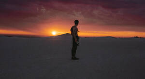 Ein Mann betrachtet den Sonnenuntergang in der Wüste, während er trainiert. Ein männlicher Sportler macht eine Pause vom Training auf den Sanddünen. - JLPSF20138
