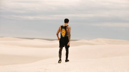 Rückansicht eines fitten jungen Mannes mit Trinkrucksack, der über Sanddünen läuft. Männlicher Sportler beim Laufen in der Wüste. - JLPSF20145