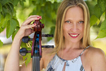Glückliche junge Frau mit Cello an einem Baum stehend - DLTSF03360