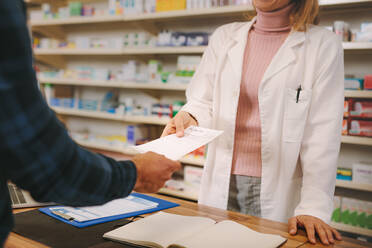 Ein Kunde übergibt ein medizinisches Rezept an eine Apothekerin, die hinter dem Tresen steht. Eine Apothekerin nimmt ein Rezept von einem Kunden in einer Drogerie entgegen. Geschnittene Aufnahme. - JLPSF20153