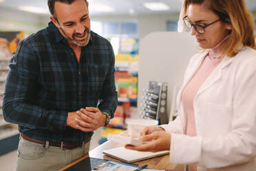 Female pharmacist holding medicine box giving advice to customer in chemist shop. Pharmacist suggesting medical drug to buyer in pharmacy drugstore. - JLPSF20156