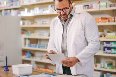 Male pharmacist reading prescription at checkout counter. Mature male chemist working in drug store. - JLPSF20157