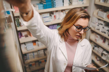 Weibliche Apothekerin hält Rezept suchen Medizin in der Apotheke zu speichern. Chemiker Blick auf Rezept bei der Suche vorgeschriebenen Medikament auf Regalen in der Apotheke. - JLPSF20159