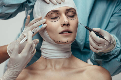 Woman wrapped in medical bandages while doctors with syringe and scalpel near her face. Female having a face lift surgery at cosmetic clinic. - JLPSF20189
