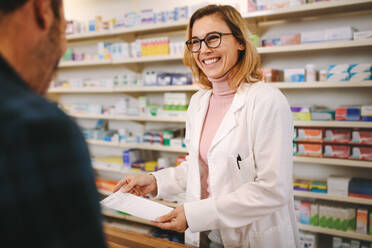 Lächelnde Apothekerin mit Rezept, die einem am Tresen stehenden Kunden hilft. Hilfreiche Apothekerin, die sich um einen männlichen Kunden kümmert, der an ihrem Schreibtisch in der Apotheke steht, sie lächelt, während sie ein Rezept erklärt. - JLPSF20232