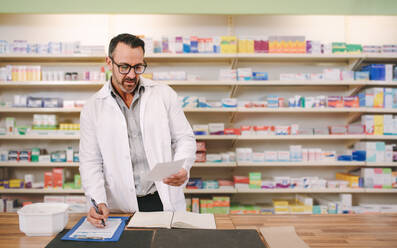 Male pharmacist writing prescription at workplace. Mature male chemist reading a prescription paper and writing on clipboard at counter. - JLPSF20237