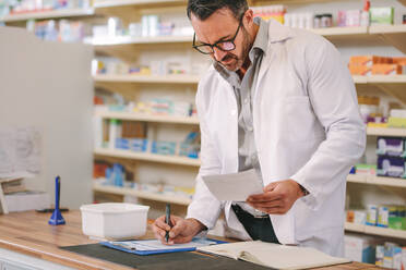 Male pharmacist writing prescription pharmacy counter. Mature druggist writing prescription on clipboard at counter. - JLPSF20238