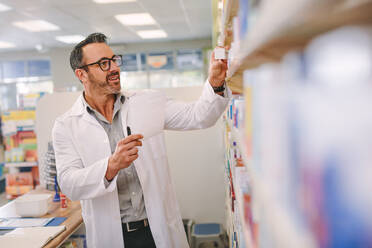 Mature chemist with a prescription searching right medicine on shelves in pharmacy. Male pharmacist holding prescription checking medicine in pharmacy. - JLPSF20240