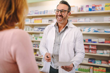 Helpful male pharmacist dealing with a woman customer standing at desk in the pharmacy. Smiling male pharmacist with prescription assisting a customer standing at the counter. - JLPSF20241