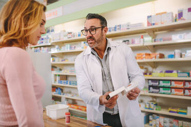 Male pharmacist holding medicine box giving advice to female customer in chemist shop. Pharmacist suggesting medical drug to buyer in drugstore. - JLPSF20243