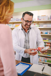 Chemist suggesting medical drug to buyer in pharmacy drugstore. Professional male pharmacist showing medicine to female customer in pharmacy. - JLPSF20244