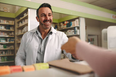Ein Kunde übergibt dem lächelnden Apotheker, der hinter dem Tresen steht, ein medizinisches Rezept. Ein Apotheker nimmt ein Rezept von einem Kunden in einer Drogerie entgegen. - JLPSF20246