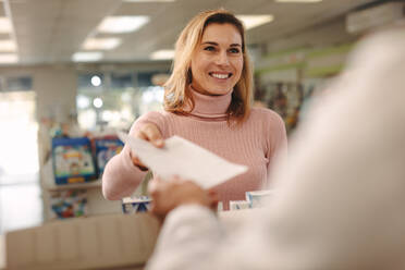 Customer giving a prescription to the chemist standing at counter. Woman standing at medical store counter and handing a prescription to pharmacist. - JLPSF20248