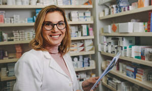 Glückliche Apothekerin mit Brille, die ein Rezept in der Hand hält und nach dem richtigen Medikament in den Regalen der Apotheke sucht. - JLPSF20257