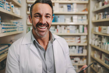 Smiling mature pharmacist with digital tablet standing by aisle in drug store. Male pharmacist working in medical store. - JLPSF20258