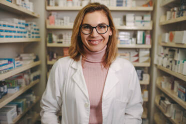 Confident female pharmacist in lab coat standing in pharmacy and looking at camera smiling. Female chemist standing in aisle of medicine racks. - JLPSF20259