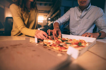Geschäftskollegen, die sich spät in der Nacht im Büro eine Pizza teilen. Ausschnitt eines Paares, das zusammen sitzend eine Pizza isst. - JLPSF20268