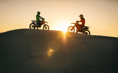 Motocross riders riding on their bikes on sand dune. Motorcycle riders on their bikes opposite to each other on a sand dune with sun in the background. - JLPSF20274