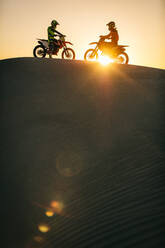 Motocross bikers sitting on their bikes on a sand dune. Motorcycle riders on their bikes standing opposite to each other on a sand dune with sun in the background. - JLPSF20276