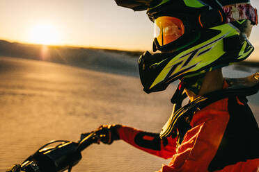 Nahaufnahme eines Motocross-Fahrers in Rennkleidung, der auf einem Motorrad in der Wüste sitzt. Seitenansicht eines Motorradfahrers mit Helm während eines Motocross-Rennens auf einer Sanddüne mit Sonne im Hintergrund - JLPSF20277