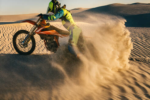 Motocross-Fahrer fährt mit seinem Motorrad auf einer Sanddüne. Motorradfahrer in Rennkleidung fährt mit seinem Motorrad die Sanddüne hinauf und hinterlässt Sandstaub. - JLPSF20280
