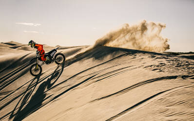 Motocross rider riding his racing bike down the sand dune. Motorbike rider in racing gear riding in the sand leaving a trail of sand dust. - JLPSF20289