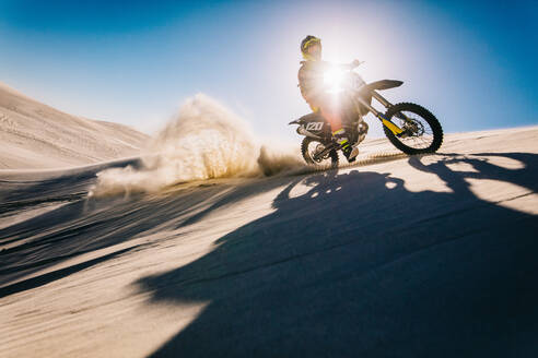Professionelle Motocross-Biker fahren sein Motorrad auf einer Sanddüne mit seinem Schatten vor. Motorradfahrer in Rennkleidung Reiten auf der Sanddüne verlassen eine Spur von Sand Staub mit Sonne im Hintergrund. - JLPSF20297