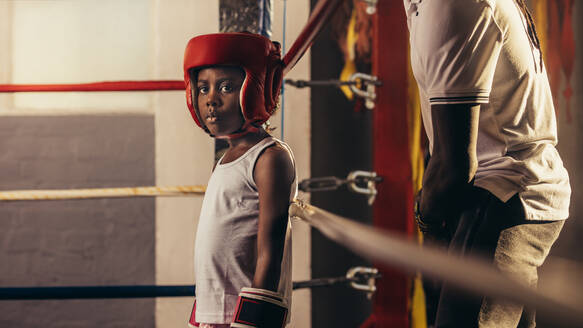 Ein Junge mit Boxhandschuhen und Kopfbedeckung steht in einer Ecke eines Boxrings. Ein kleiner Junge steht in einem Boxring mit seinem Trainer hinter ihm. - JLPSF20306