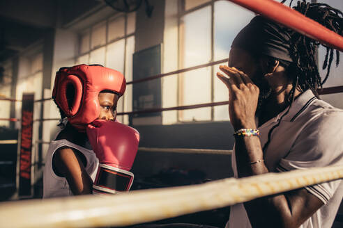 Boxtrainer gibt einem Kind in einer Boxhalle Anweisungen. Ein Kind mit Boxhandschuhen und Kopfbedeckung trainiert mit seinem Trainer in einem Boxring. - JLPSF20313