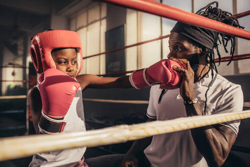 Ein Kind übt mit Boxhandschuhen und Kopfbedeckung mit seinem Trainer Schläge. Ein Boxtrainer bringt einem Kind die Schlagtechniken bei. - JLPSF20314