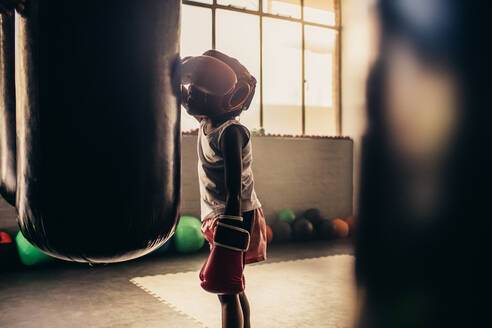 Müdes Kind mit Boxhandschuhen und Kopfbedeckung, das sich an einen Boxsack lehnt. Boxendes Kind, das nach dem Training müde dasteht. - JLPSF20321