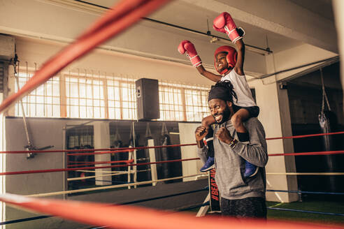 Ein Trainer trägt ein Boxerkind auf den Schultern und feiert einen Sieg. Ein Kind in Boxerausrüstung sitzt mit erhobenen Händen auf der Schulter des Trainers. - JLPSF20334