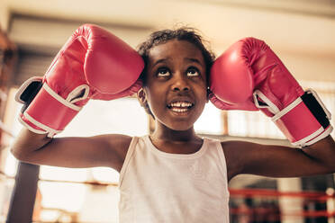 Kid wearing boxing gloves standing in a boxing ring touching her head with gloves and looking up. - JLPSF20335